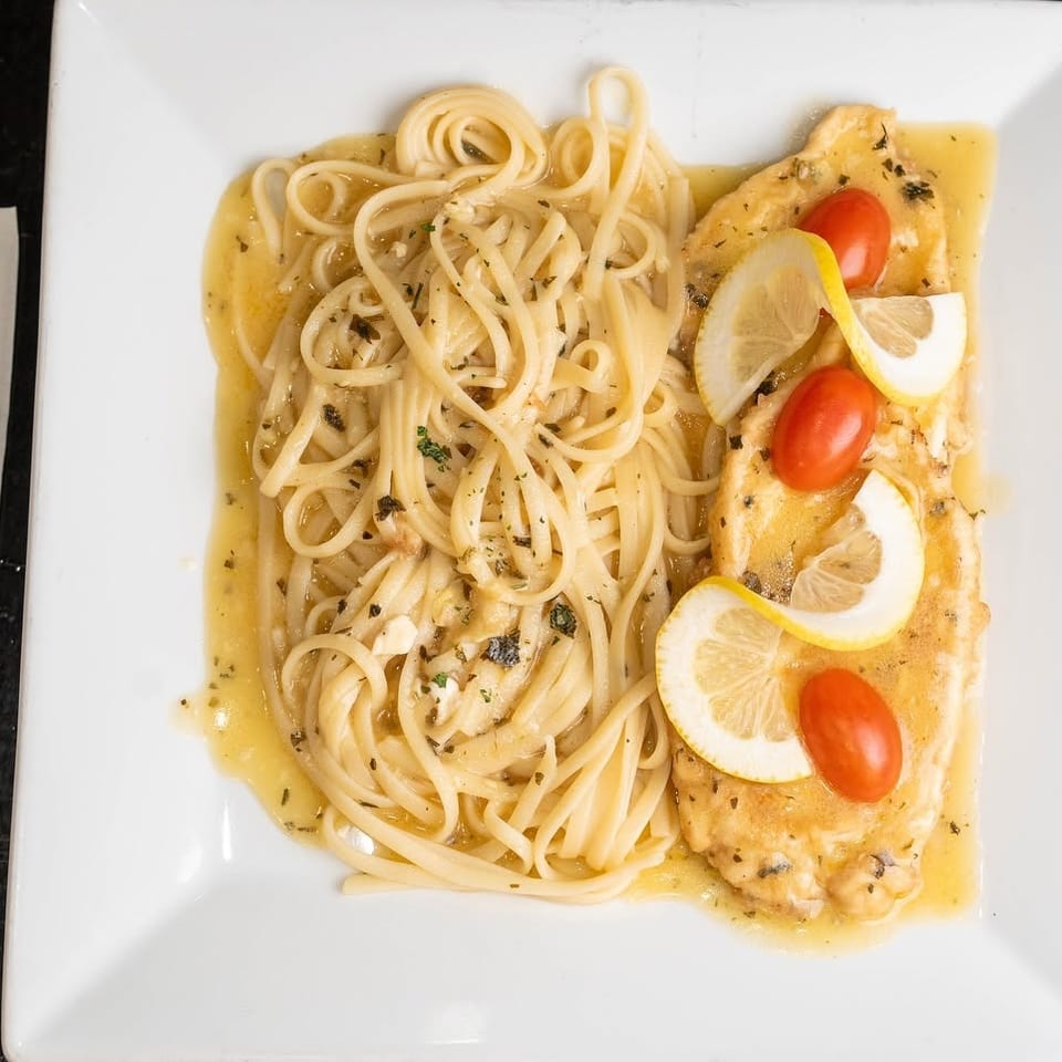 Linguine and grilled chicken breast in lemon herb sauce, garnished with lemon slices and cherry tomatoes on a white plate.
