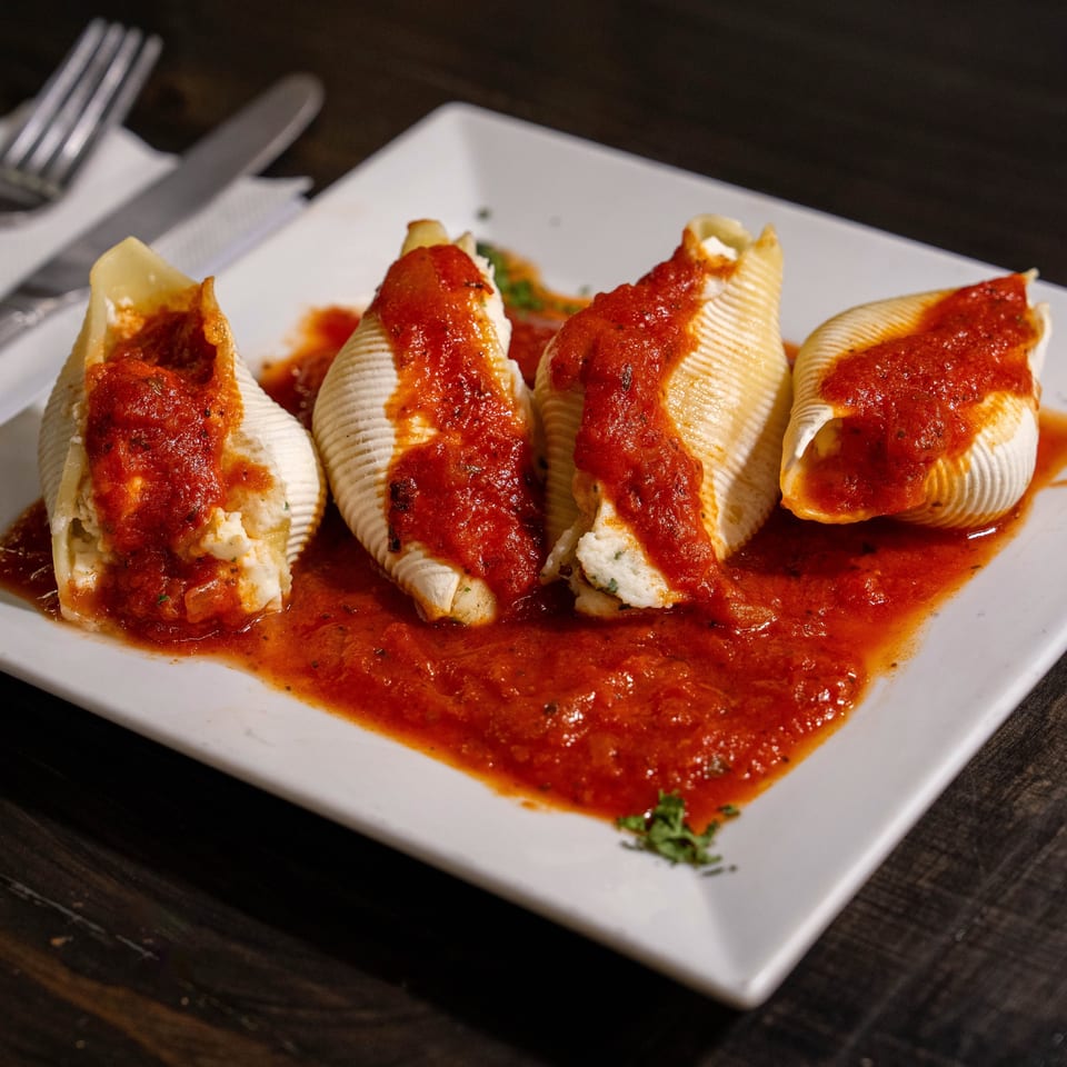 Four stuffed pasta shells topped with tomato sauce and ricotta, served on a white plate with utensils.