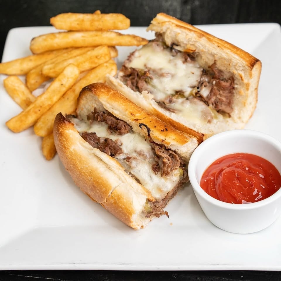 Cheesesteak sandwich halves with melted cheese, golden fries, and ketchup on a white plate.
