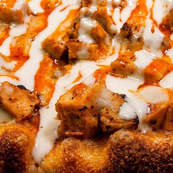 Close-up of a pizza topped with chicken pieces, white sauce, and orange hot sauce drizzle.