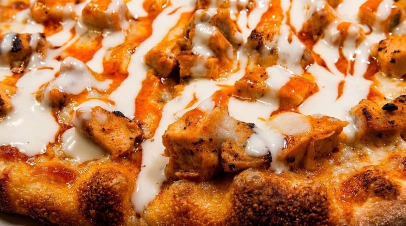 Close-up of a pizza topped with chicken pieces, white sauce, and orange hot sauce drizzle.